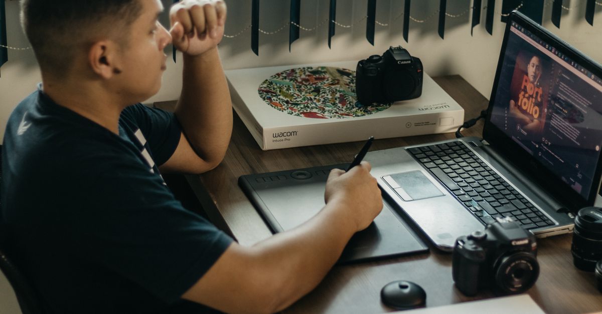 A photographer working at a creative setup with a laptop and Wacom tablet, focused on editing photos indoors.